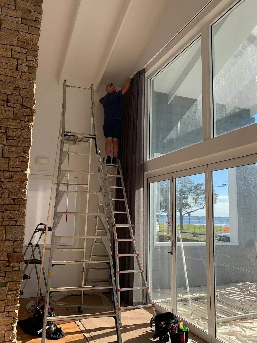 Installer on a ladder fitting a curtain track above a tall living room window